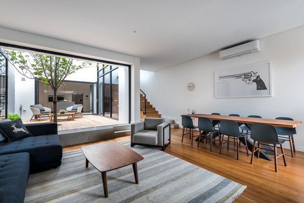 Bright open-plan living and dining area with a contemporary minimalist interior. The room features a dark sectional sofa, grey armchair, timber coffee table, long timber dining table with upholstered chairs, pale striped rug, white walls, timber flooring, and large glazed openings connecting to a landscaped courtyard. A stairway and split-system air conditioner are visible, along with framed wall art and a small wall clock.