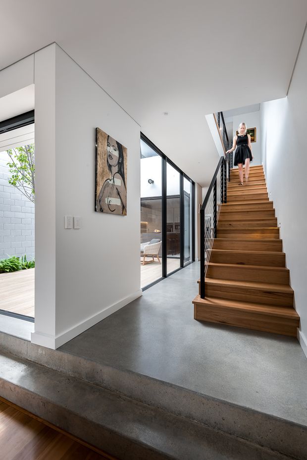 A bright, modern residential hallway centered on a timber staircase with black metal balustrades. The polished concrete floor continues across split levels, while full-height black-framed glazing opens toward a light-filled courtyard and adjacent sitting area. White walls and ceilings create a clean backdrop, with a framed artwork mounted beside the stair. A person is visible on the upper stairs.
