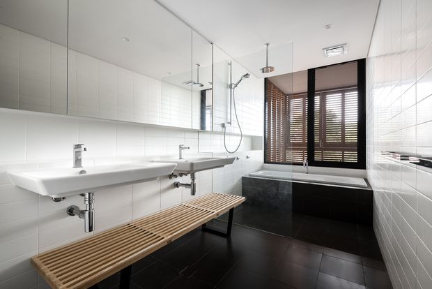 Contemporary bathroom with two wall-mounted white basins, exposed chrome plumbing, a mirrored wall, walk-in glass shower, freestanding bath, dark tiled floor and bath surround, and timber Venetian blinds at the window. A slatted timber bench runs beneath the basins. The room is bright, clean and highly functional, with strong black-and-white contrast softened by timber details.