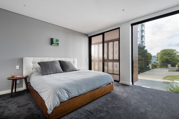 A contemporary bedroom with a low-profile timber bed featuring a padded white headboard, grey bedding and charcoal cushions. The room has wall-to-wall dark grey carpet, smooth light grey walls, a white ceiling with recessed downlights, and full-height black-framed glazing with timber louvred shutters. A small round timber side table and compact artwork provide minimal furnishings, while the glazing overlooks a leafy suburban streetscape.