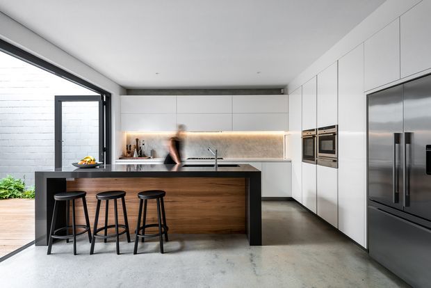 A contemporary open-plan kitchen with a large island featuring a dark stone or composite benchtop, a warm timber-faced base, and three black bar stools. Full-height white integrated cabinetry lines the rear and right walls, incorporating double ovens and a large stainless-steel refrigerator. A linear illuminated splashback and under-cabinet lighting provide a warm accent, while polished concrete flooring, dark-framed glazing, and a partially open door create a clean, minimalist setting.