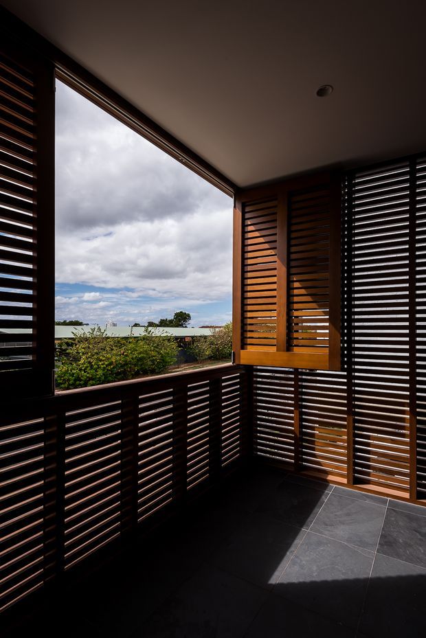 A sheltered balcony or terrace enclosed with dark timber louvred screens and balustrades. Large openings frame a cloudy sky, distant low rooftops, and planted greenery. The space has a smooth light ceiling, dark large-format floor tiles, and strong contrast between the warm timber screens and the shadowed interior.