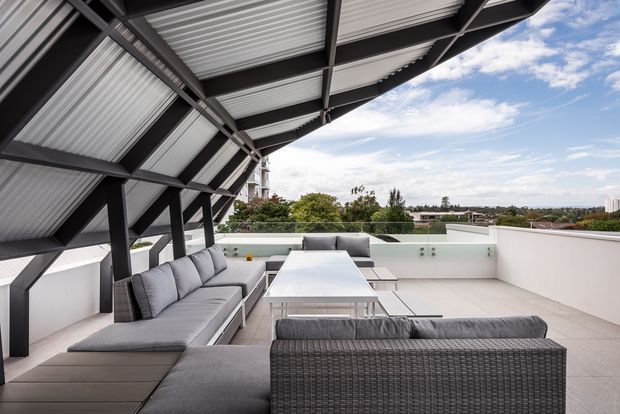 A spacious rooftop terrace with a contemporary outdoor lounge beneath a dramatic sloping corrugated metal canopy. The terrace features grey sectional seating, a low rectangular outdoor table, pale tiled flooring, white rendered parapet walls, and dark exposed steel framing. Trees, rooftops, and a distant urban skyline are visible beyond the terrace.