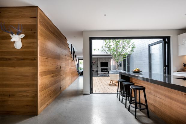 A contemporary open-plan kitchen and living area with a long timber-clad wall of integrated joinery on the left and a substantial kitchen island on the right. The island has a dark stone benchtop, warm timber-panelled base, and three black bar stools. Large black-framed glazed doors open to a timber deck and a courtyard-like living space beyond. The interior has polished concrete flooring, white walls and ceiling, recessed lighting, and a restrained natural-material palette.
