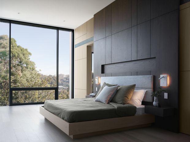 Contemporary bedroom with a low-profile floating timber bed, layered neutral bedding, and a dark timber feature wall with integrated headboard and bedside shelving. Full-height black-framed glazing opens to leafy views, while warm wall-mounted bedside lights add contrast against the dark joinery.