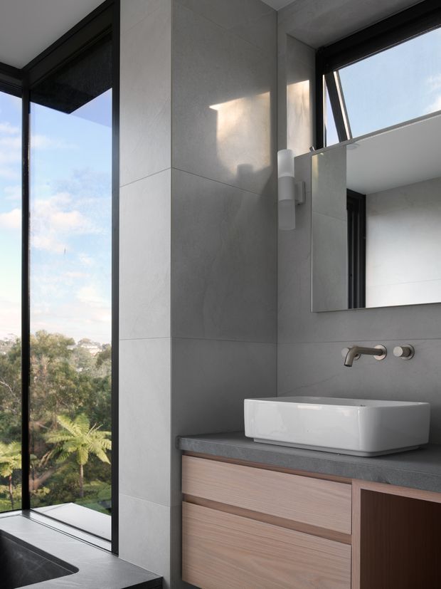 Contemporary bathroom vanity in a bright, refined space, featuring large-format grey wall tiles, a white vessel basin, wall-mounted brushed metal tapware, timber-look floating cabinetry, a dark benchtop, frameless mirror, and black-framed floor-to-ceiling glazing overlooking greenery.