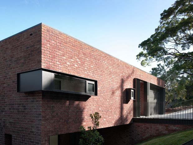 Exterior view of a minimalist single-storey brick building with a long, low rectangular form, flat roofline, dark-framed projecting windows, and a recessed glazed opening with vertical metal balustrades. The building sits on a slightly sloped landscaped site beneath mature trees.