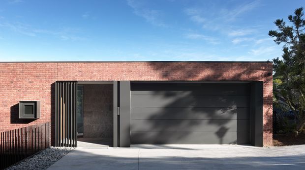 Exterior view of a low, modern brick-clad building with a broad dark sectional garage door, narrow vertical metal screening at the entry, and a small square window. The flat-roofed facade is strongly horizontal and set against a clear blue sky, with tree shadows cast across the brick and garage door.
