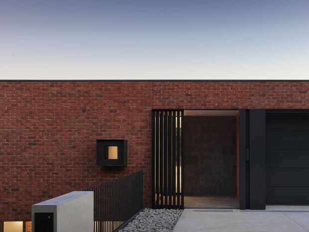 Contemporary single-storey brick facade with a flat roofline, dark metal-framed entry, vertical screening, recessed doorway, and integrated dark garage door. A concrete path and gravel bed lead to the entrance, with a small square wall light set into the brickwork.
