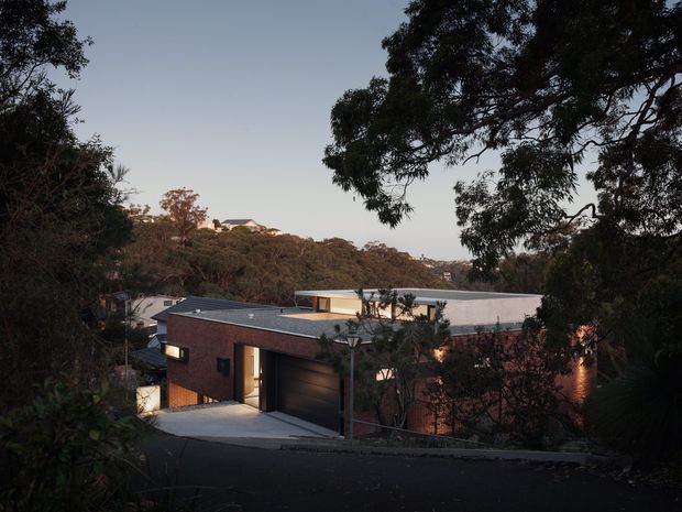 Exterior dusk view of a modern low-rise brick house on a sloping, wooded suburban site. The home has a dark metal-looking low-pitched roof, broad flat-roofed sections, dark-framed openings, warm interior lighting, and an integrated double garage accessed from a sloping driveway. Dense trees frame the foreground, with additional homes visible across the distant hillside.