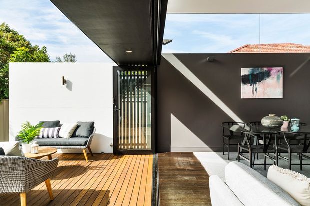 A bright contemporary indoor-outdoor living and dining area connected by wide sliding glass doors. The space features a timber deck, a dark timber-toned interior floor, white and charcoal walls, black-framed glazing, a compact outdoor lounge with sofa and armchair, and an adjacent dining setting under a covered roof. Strong sunlight creates a crisp, relaxed entertaining space with tropical greenery visible beyond.