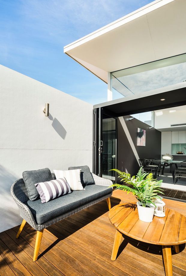 Bright architectural photograph of a modern outdoor terrace with timber decking, a grey woven outdoor sofa, and a round timber coffee table with potted greenery. Large open black-framed glazing connects the terrace to a contemporary dining area, while clean white walls, a projecting flat roof, and strong sunlight create a crisp indoor-outdoor composition.