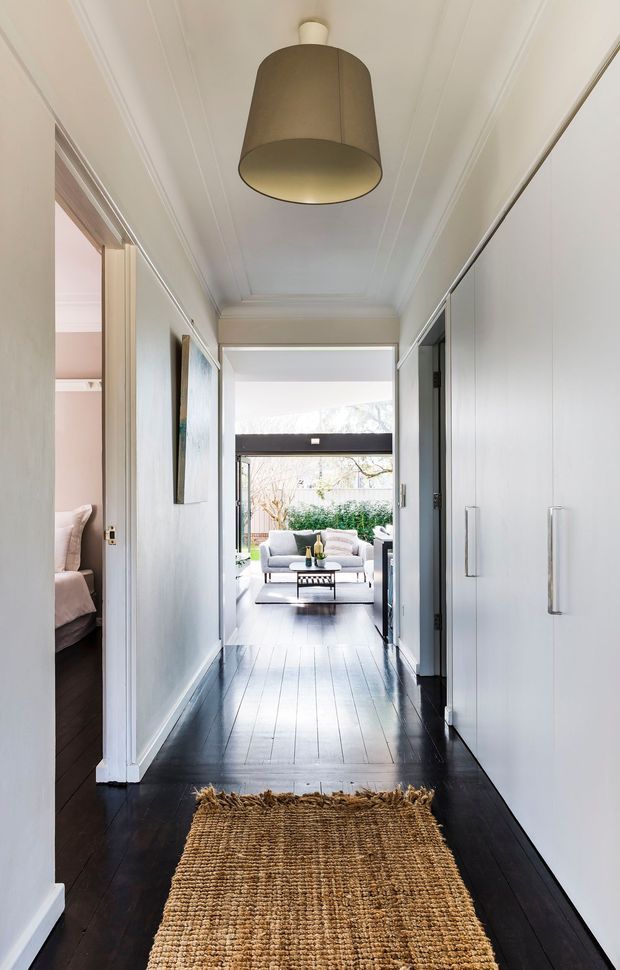 A bright, narrow residential hallway viewed toward an open living area and garden beyond. The corridor has dark timber floorboards, white walls and cabinetry with long vertical handles, decorative white ceiling mouldings, a large drum pendant light, and a natural woven runner. An open bedroom doorway is visible on the left, while a light-filled living room with seating is at the far end.