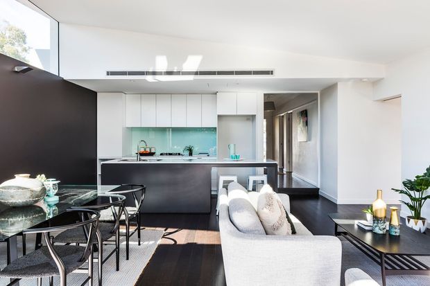 Bright open-plan contemporary interior showing a compact kitchen behind a dark island, a dining area with a round glass table and black chairs, and a light upholstered sofa in the foreground. White cabinetry and walls contrast with dark timber flooring and a turquoise glass-tile splashback, with large glazing bringing in strong daylight.