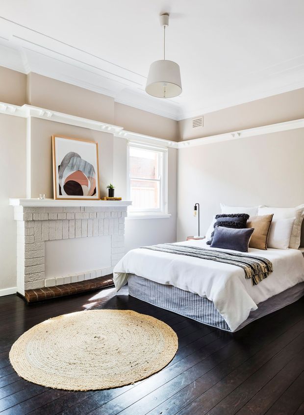 Bright, refined bedroom with a white-painted brick fireplace, dark timber floors, beige walls, white decorative cornices and picture rails, and a large upholstered bed dressed in layered neutral bedding. A woven round rug, abstract artwork, pendant light and soft natural light complete the calm interior.