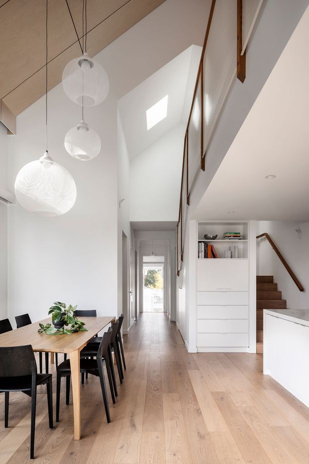 A bright, double-height open-plan dining and circulation space with pale timber flooring, white walls, and a warm timber-lined ceiling. A rectangular timber dining table with black chairs sits beneath three suspended white spherical pendant lights. The central hallway leads toward a glazed rear doorway, while a white built-in storage unit with open shelving and drawers separates the hall from a timber staircase. A mezzanine-level opening with slim timber balustrade detailing overlooks the space. The overall design is clean, airy, and contemporary with natural timber accents.