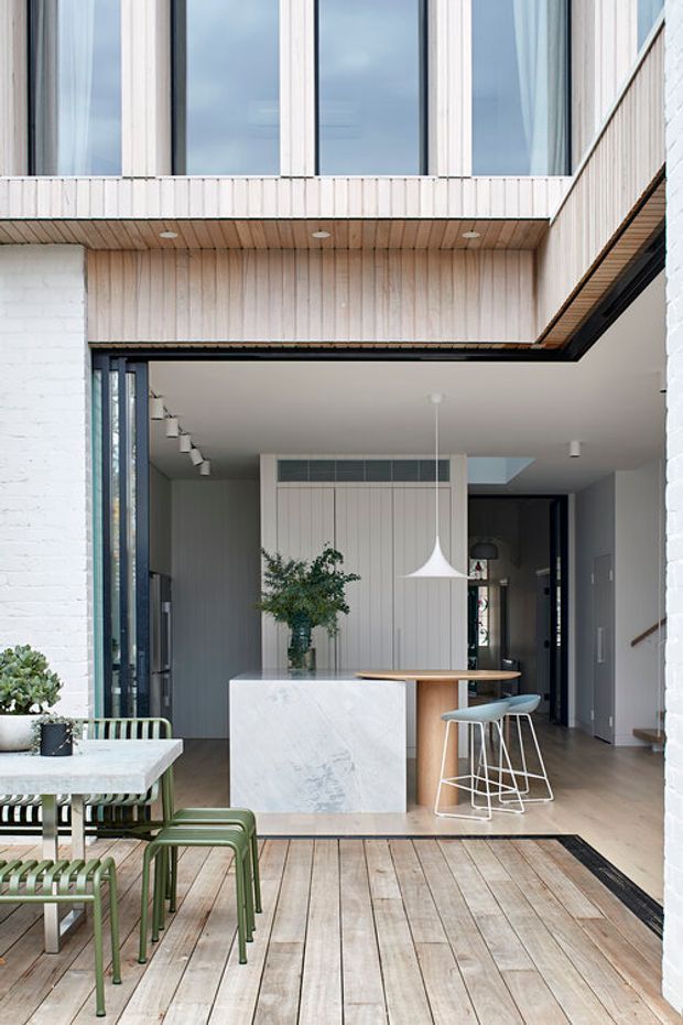 A bright contemporary indoor-outdoor living space viewed from a timber deck. The covered opening frames a compact kitchen with a white marble island, timber breakfast counter, pale vertical wall panelling, pendant lighting, and bar stools. An outdoor dining setting with green-framed chairs sits in the foreground. White-painted brick walls, extensive glazing, black-framed doors, and timber soffit lining define the architecture.