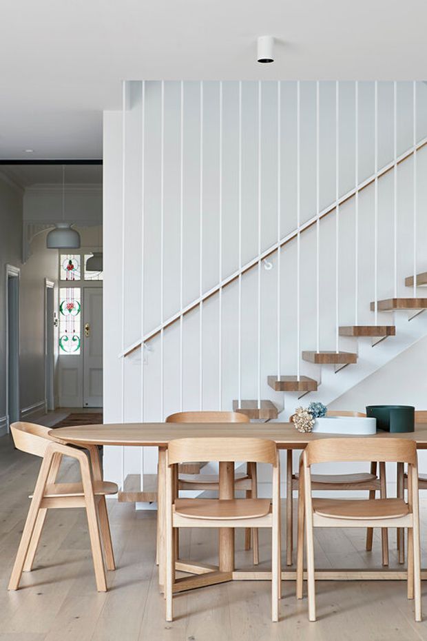 Bright, refined dining area centred on a light timber dining table and matching chairs. A sculptural open staircase with floating timber treads, slender white vertical balusters, and a white panelled stair wall dominates the background. The room has pale timber flooring, white walls and ceiling, minimalist cylindrical lighting, and a glimpse of a traditional entry hall with decorative glazed doors beyond.
