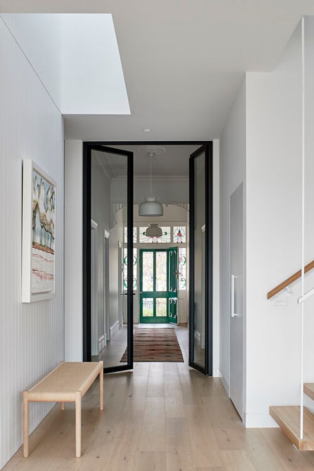 A bright residential hallway and entryway with pale timber flooring, white walls, and a large skylight. Black-framed glazed double doors open toward a green decorative glass front door at the end of the corridor. A simple timber bench sits along the left wall, while a staircase with a timber handrail rises on the right. The space combines crisp contemporary detailing with traditional decorative glazing and ceiling mouldings.