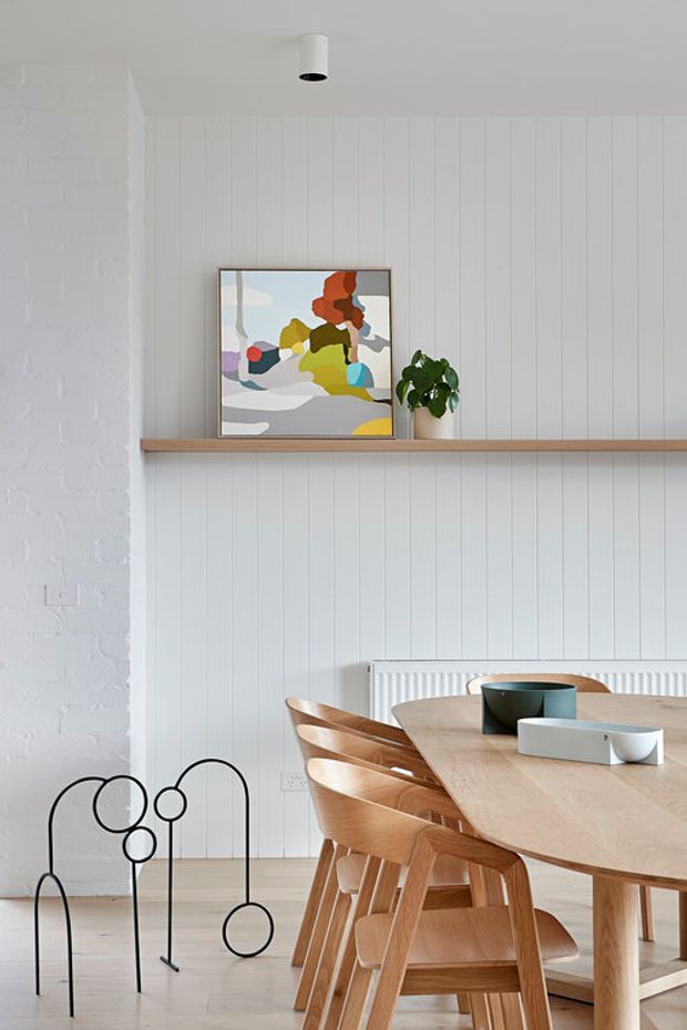 Bright dining area with a long light-wood dining table and matching wooden chairs, set against white vertical timber wall panelling and a white painted brick wall. A slim timber display shelf holds a framed abstract artwork and potted plant. The room includes a white radiator, pale flooring, and a minimalist black sculptural floor light.