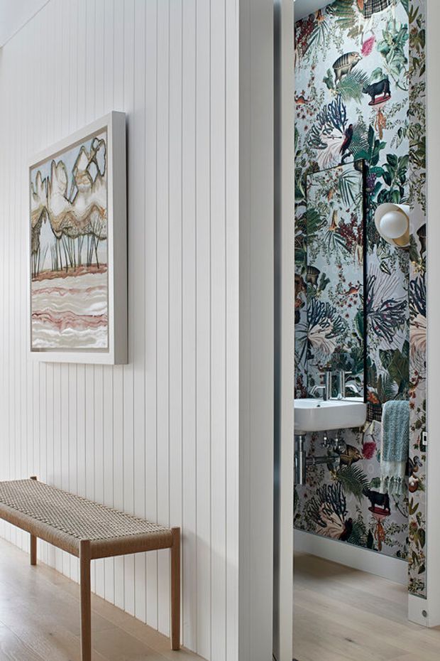 A bright transitional hallway or entry area with white vertical wall panelling, pale timber flooring, a woven timber bench, and framed artwork. Through the doorway is a compact bathroom featuring vivid botanical and bird-patterned wallpaper, a wall-mounted white basin, chrome tapware, and a textured blue towel.