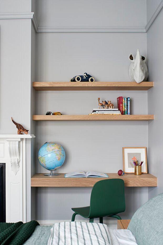 A refined children's bedroom with a built-in study nook, featuring three floating light-wood shelves against a soft grey wall. The shelves display books, toys, a globe, framed artwork and stationery, with a green desk chair tucked beneath the lowest shelf. Part of a bed with pale blue-green bedding is visible in the foreground, while a white decorative fireplace surround with classic moulding appears at left. The room combines traditional architectural detailing with playful, contemporary furnishings.