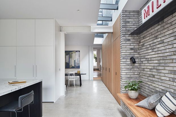Bright contemporary open-plan interior showing a kitchen at left, a long hallway with extensive timber built-in cabinetry, a dining area in the centre, and a brick feature wall with built-in bench seating at right. Polished concrete flooring, white cabinetry, skylights, and a restrained neutral palette create a clean, modern atmosphere.