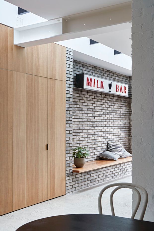 Bright contemporary interior featuring tall light-wood built-in cabinetry, an exposed white-painted brick feature wall with a timber bench seat, and a prominent “MILK BAR” sign. A round dark dining table and curved chairs occupy the foreground. Overhead skylights and layered ceiling planes bring in abundant natural light.