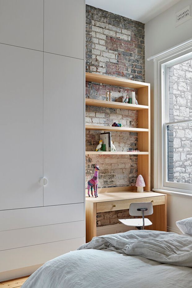 A compact, bright bedroom with a built-in pale cabinetry wall, integrated open timber shelving and a small study desk. The shelving is set against an exposed, weathered brick wall, while a large traditional sash window brings in natural light. A bed with soft light bedding occupies the foreground, and children's toys and a small chair suggest a youthful bedroom or study nook.