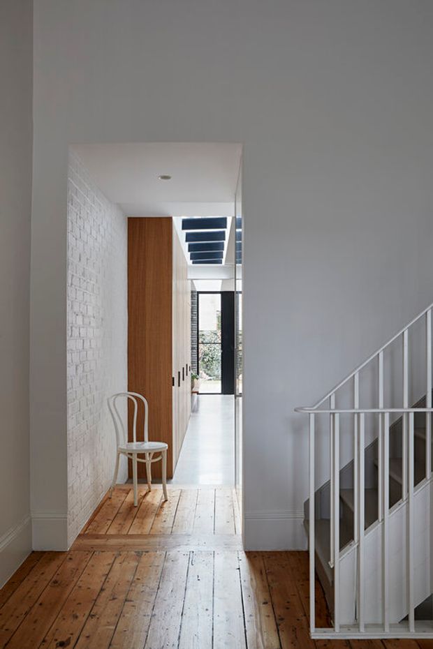 A bright, narrow residential hallway with original timber floorboards, a white-painted brick wall, and a white staircase with slim balustrades in the foreground. A minimalist corridor extends toward a glazed rear door, with tall timber cabinetry and a simple bentwood chair along the passage. The restrained palette and clean lines create a calm, contemporary interior with preserved traditional fabric.