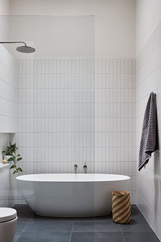 A bright, minimalist bathroom featuring a freestanding white oval bathtub beneath a wall-mounted rain shower. White vertically arranged wall tiles extend around the bath, while large dark grey floor tiles provide contrast. A partial toilet, small plant, woven basket, and patterned towel are visible.