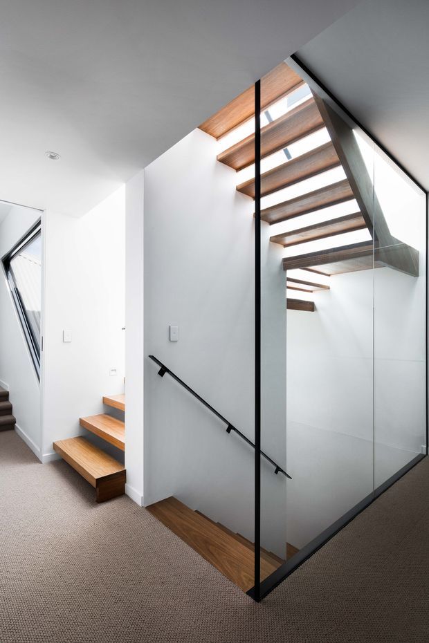 Bright contemporary stairwell with floating timber treads and exposed timber ceiling beams, enclosed by a tall frameless glass balustrade with slim black metal framing. White walls and ceilings contrast with the warm wood, while carpeted flooring continues through the landing. A diagonal black handrail and skylight-like glazing create a light-filled, vertically layered space.