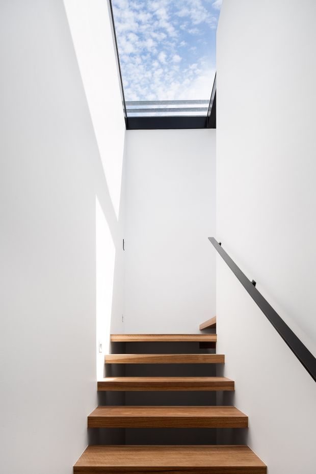 Minimalist interior stairway with floating timber treads, dark recessed risers, crisp white walls, and a slim black handrail. A large roof skylight brings in daylight and reveals a partly cloudy sky above the stairwell.