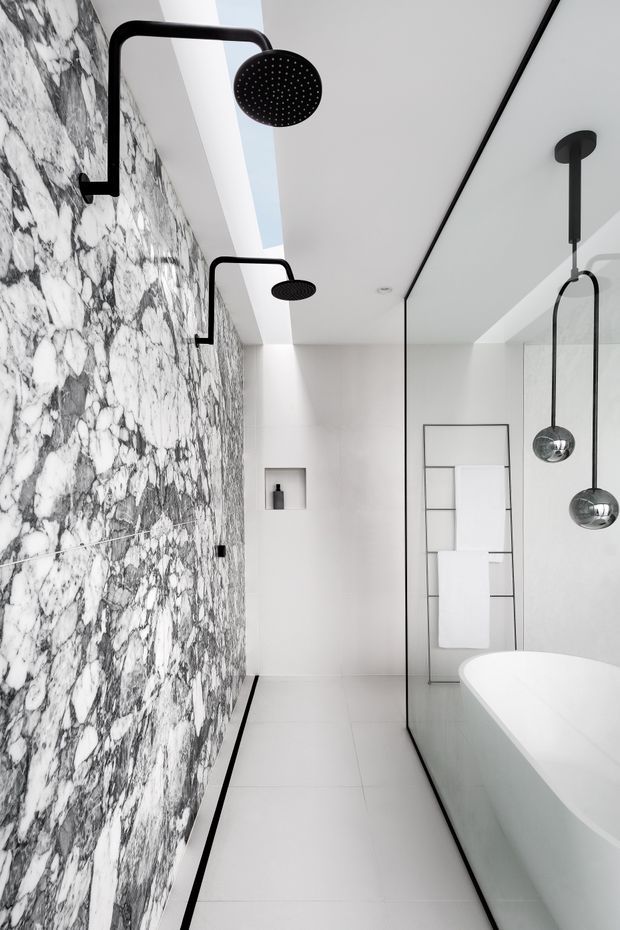A refined contemporary bathroom featuring a dramatic full-height white-and-grey marble feature wall with matte black shower fittings. A frameless glass shower screen separates the shower area from a freestanding white bathtub, complemented by black pendant-style fixtures and a minimalist heated towel ladder. The palette is predominantly white, light grey, black, and marble veining, creating a luxurious high-contrast interior.