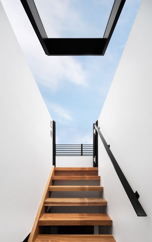 A narrow modern stairway rises between crisp white walls, with floating warm timber treads, dark structural elements, and a black handrail. The stairwell opens to a bright blue sky through a large overhead skylight and a small upper-level opening with horizontal metal balustrading.