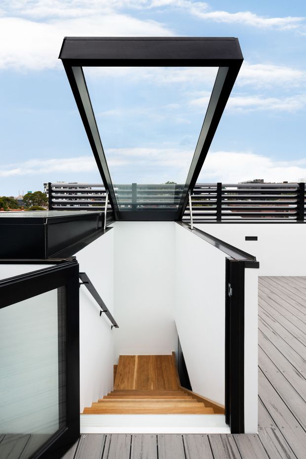 A modern rooftop terrace with an open stairwell descending into the building. The stair is framed by crisp white walls, warm timber treads and flooring, black metal handrails and balustrades, and large angular glazed skylight elements. A grey timber deck surrounds the opening, with horizontal black screening and urban apartment buildings visible beyond.