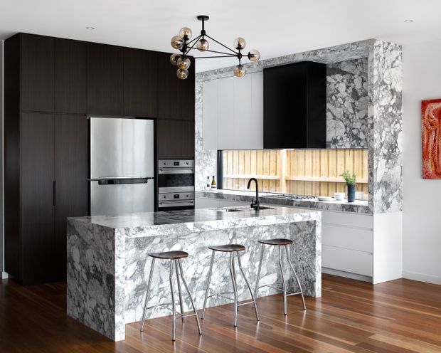 A refined contemporary kitchen featuring a large marble-clad island with seating for three, dark full-height cabinetry, stainless-steel appliances, white lower cabinetry, and a marble-framed cooking and display wall. A sculptural globe pendant light hangs above the island, with warm timber flooring and a small red artwork adding contrast.