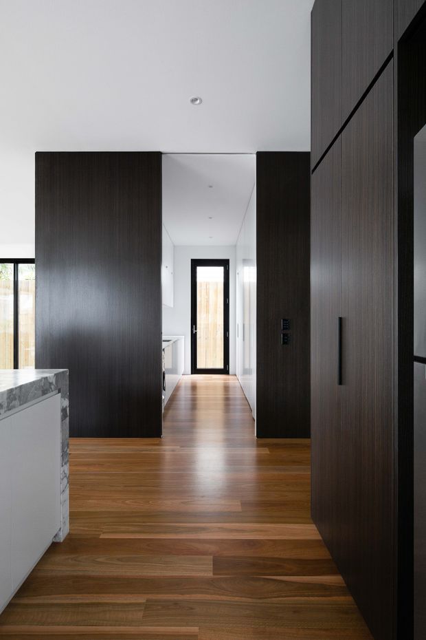 A contemporary interior hallway viewed from an open-plan kitchen area, with rich timber flooring, tall dark timber built-in cabinetry and partition walls, white walls and ceiling, and a black-framed glazed door at the end of the corridor. A marble-look kitchen island is partially visible in the foreground. The space is clean-lined, minimalist, and strongly defined by the contrast between dark joinery and warm wood flooring.