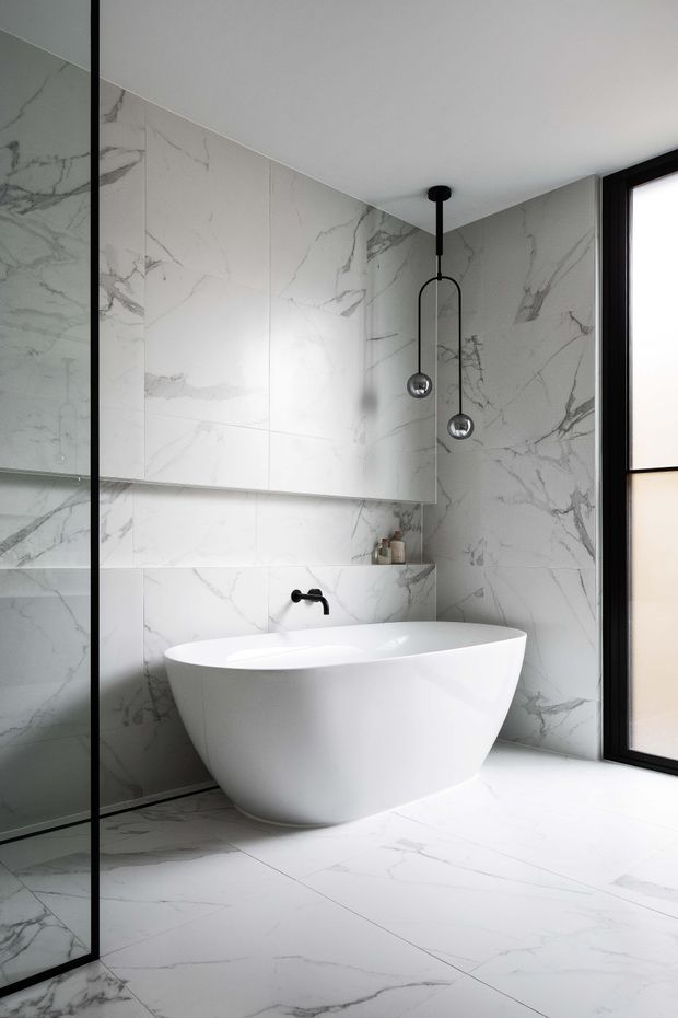 A bright, refined contemporary bathroom featuring a freestanding white oval bathtub, wall-mounted black tapware, large white marble-look tiles with grey veining on the walls and floor, a frameless black-edged glass shower screen at left, and a tall black-framed window. Minimal black pendant lights hang above the bath.