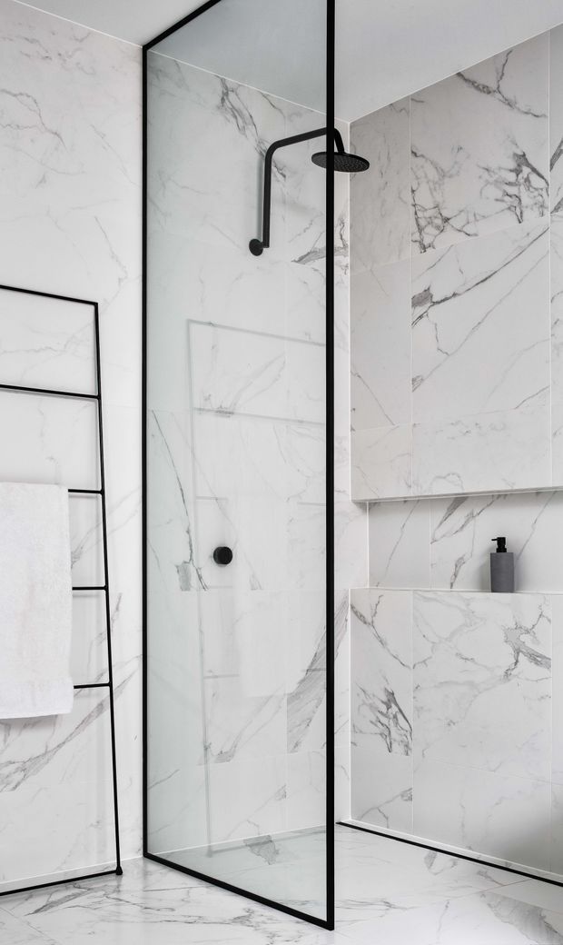 A bright, minimalist bathroom clad in large white marble-look tiles with prominent grey veining. The room features a frameless glass shower enclosure with slender black metal framing, a matte black rainfall showerhead and controls, a recessed tiled niche, a wall-mounted mirrored cabinet, and a black ladder-style heated towel rail with a white towel.