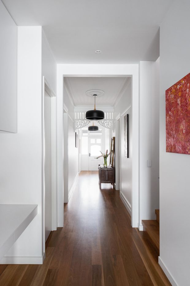 A bright, narrow residential hallway viewed from an adjoining interior space, with polished dark timber flooring, crisp white walls and trim, ornate ceiling mouldings, decorative fretwork at the far end, and black pendant lights. A console table with floral decor sits near the entry door, while a staircase and vivid red artwork are visible on the right.