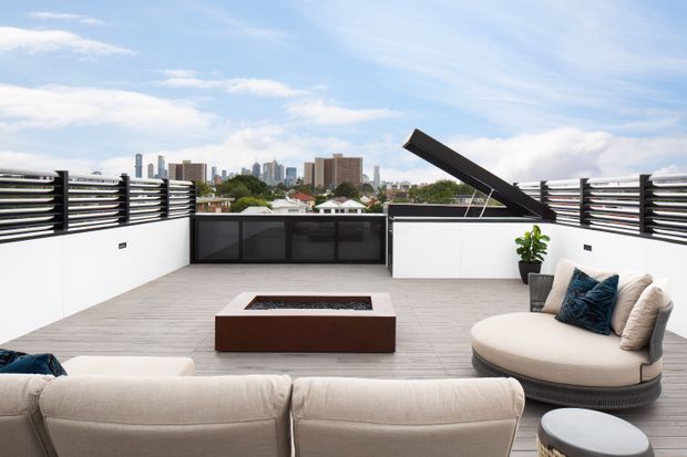 A spacious rooftop terrace overlooking a dense city skyline. The terrace features pale grey decking, white rendered privacy walls, black horizontal louvred balustrade screens, a central rectangular built-in fire pit, and a large curved outdoor sofa arrangement with cushions. A raised dark hinged panel or awning is open along the far edge, with a potted plant adding greenery. The overall design is contemporary and refined with a bright urban outdoor-living atmosphere.