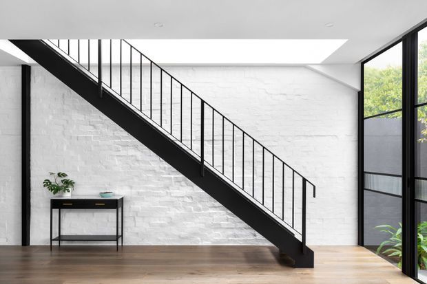 Bright minimalist interior featuring a black steel open-riser staircase with slender vertical balustrades, set against a white-painted exposed brick wall. The room has warm timber flooring, a slim black console table with a plant, a large skylight, and floor-to-ceiling black-framed glazing on the right.