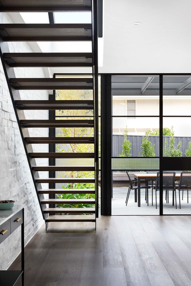 A contemporary interior featuring an open-riser staircase with dark timber treads and slim black metal supports beside a white-painted brick wall. Pale timber flooring extends toward full-height black-framed glazing and glazed doors opening to a paved outdoor dining terrace. The space is bright and minimalist, with strong indoor–outdoor visual connection and visible built-in cabinetry at the lower left.