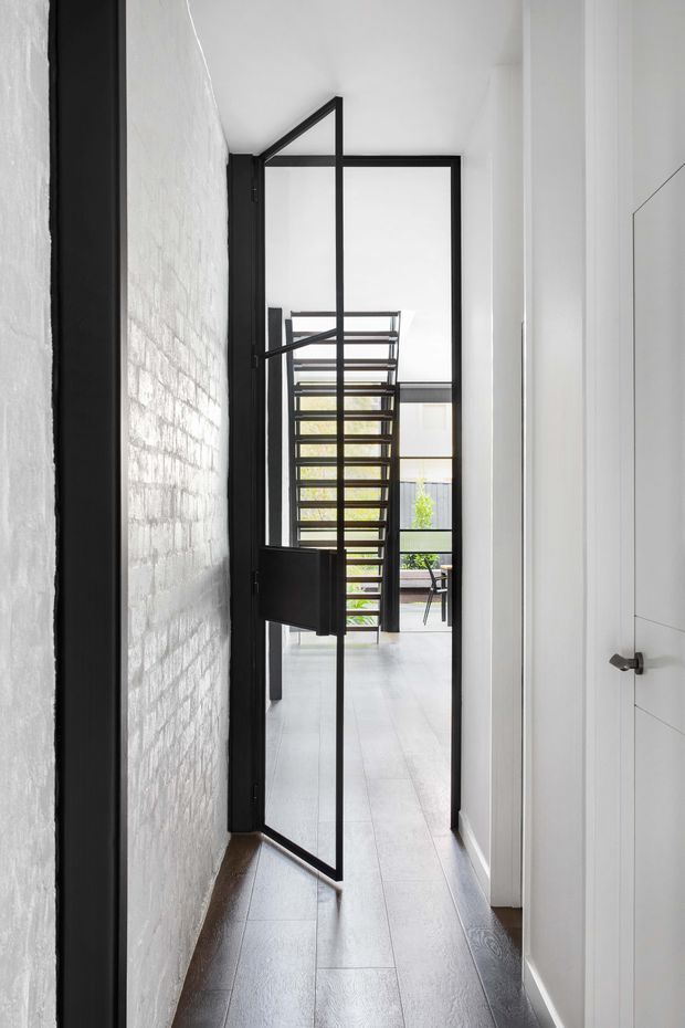 A narrow, bright interior passage with white painted brickwork on the left, white walls and a dark timber-look floor. A distinctive black steel-framed glazed door stands partly open, leading toward a light-filled living or courtyard area with a contemporary open-riser staircase and outdoor greenery visible beyond. The space has a clean, minimalist industrial character with strong black-and-white contrast.