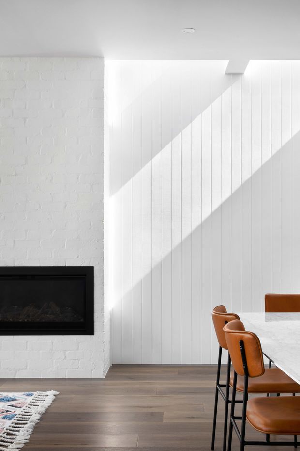 Bright contemporary dining area with a black built-in fireplace set into a white-painted brick wall, adjacent to a white vertical-panel feature wall. A pale stone dining table with warm tan leather and black metal chairs occupies the right side, while strong sunlight creates diagonal shadows across the wall. The room has dark timber-look flooring and a partially visible patterned rug.