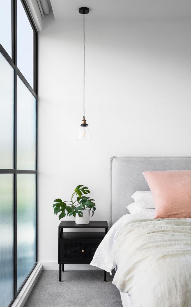 Bright, minimalist bedroom interior with a grey upholstered bed, white bedding, and a blush-pink accent pillow. A black bedside table with perforated-front storage sits beside a potted monstera plant. Full-height frosted glazing with dark metal framing occupies the left side, while a simple pendant light hangs from the white ceiling. The room has a clean, contemporary feel with soft neutral tones and strong natural light.