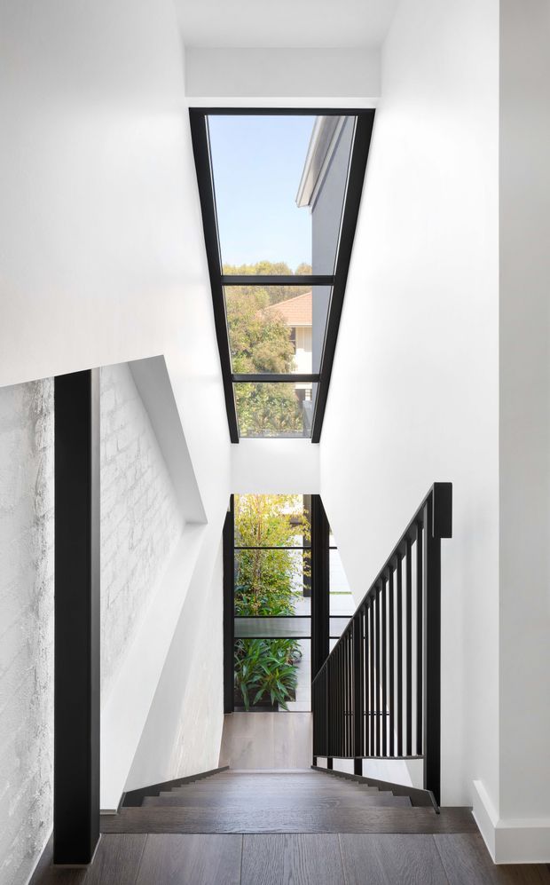 A bright modern stairway with dark timber or engineered-wood flooring, white walls, black metal balustrading and black-framed glazing. The stairwell is vertically open and naturally lit by a tall overhead skylight and full-height windows, with greenery and neighbouring residential buildings visible outside.