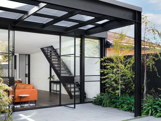 A contemporary glazed indoor-outdoor extension or courtyard viewed from a paved terrace. Black steel framing supports a translucent roof with exposed beams, while large black-framed glass doors open to a bright interior containing an orange sofa, dark timber flooring, and a sculptural staircase. Lush planting borders a dark exterior wall, with an older tiled roof visible beyond.
