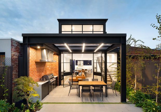 A contemporary rear extension opening onto a landscaped outdoor entertaining area. The black-framed glazed pavilion has a raised clerestory-like upper window band, exposed dark structural framing, linear strip lighting, a brick feature wall, built-in outdoor kitchen with barbecue, and a dining table beneath a covered terrace. Interior living and kitchen spaces are visible through the glazing.